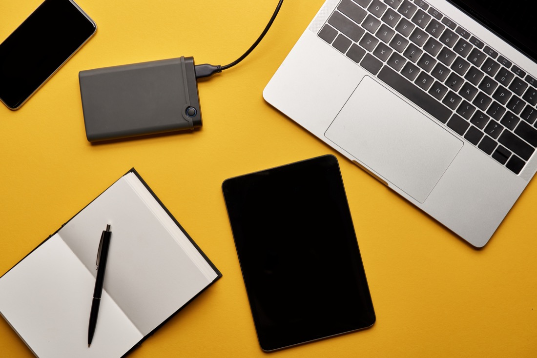 Laptop, tablet, phone, power bank, and notebook on a yellow table.