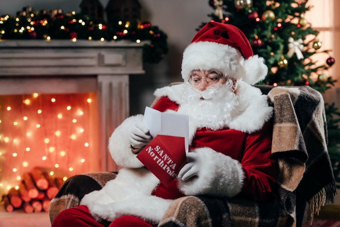 Santa sitting on a chair beside a fireplace surrounded by Christmas decor.