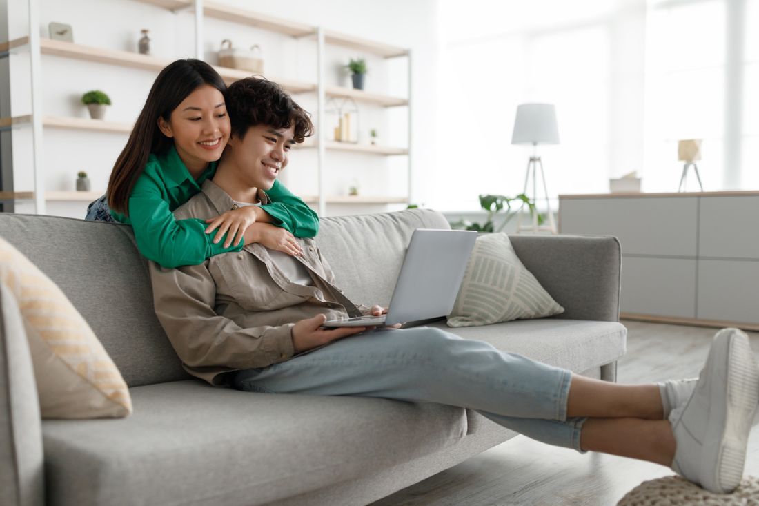 Young asian woman hugging her boyfriend who is sitting on a sofa and using a laptop.