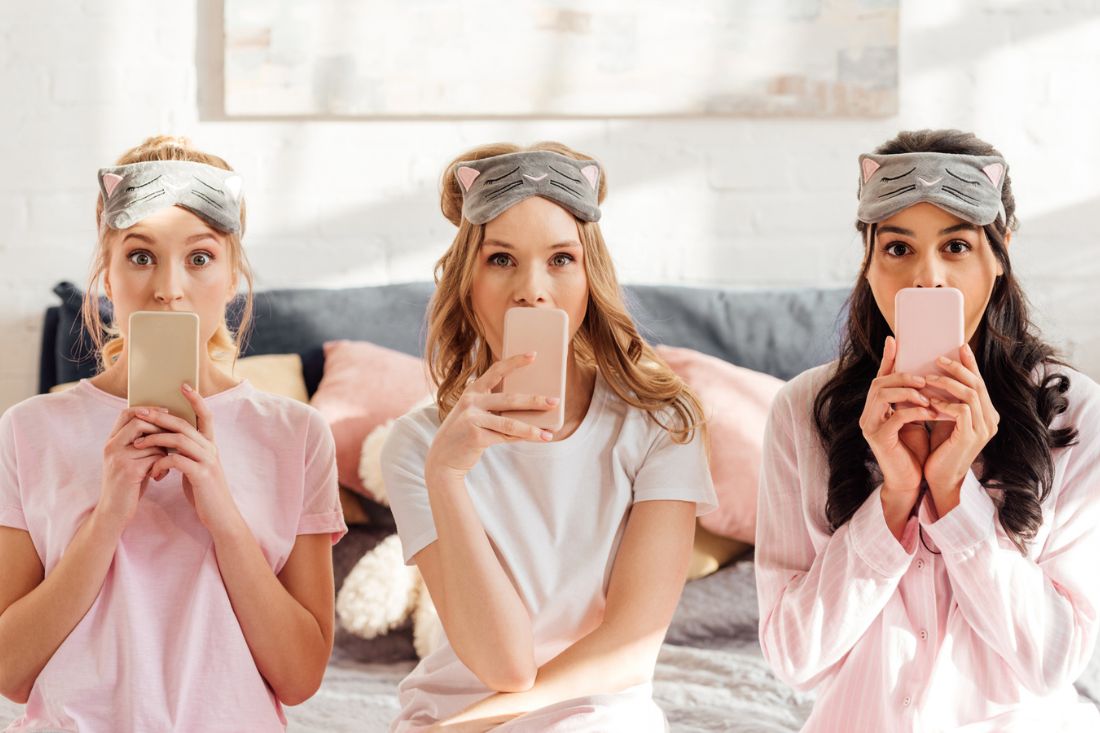 Multicultural girls in pajamas and sleeping masks sitting and covering mouths with phone.