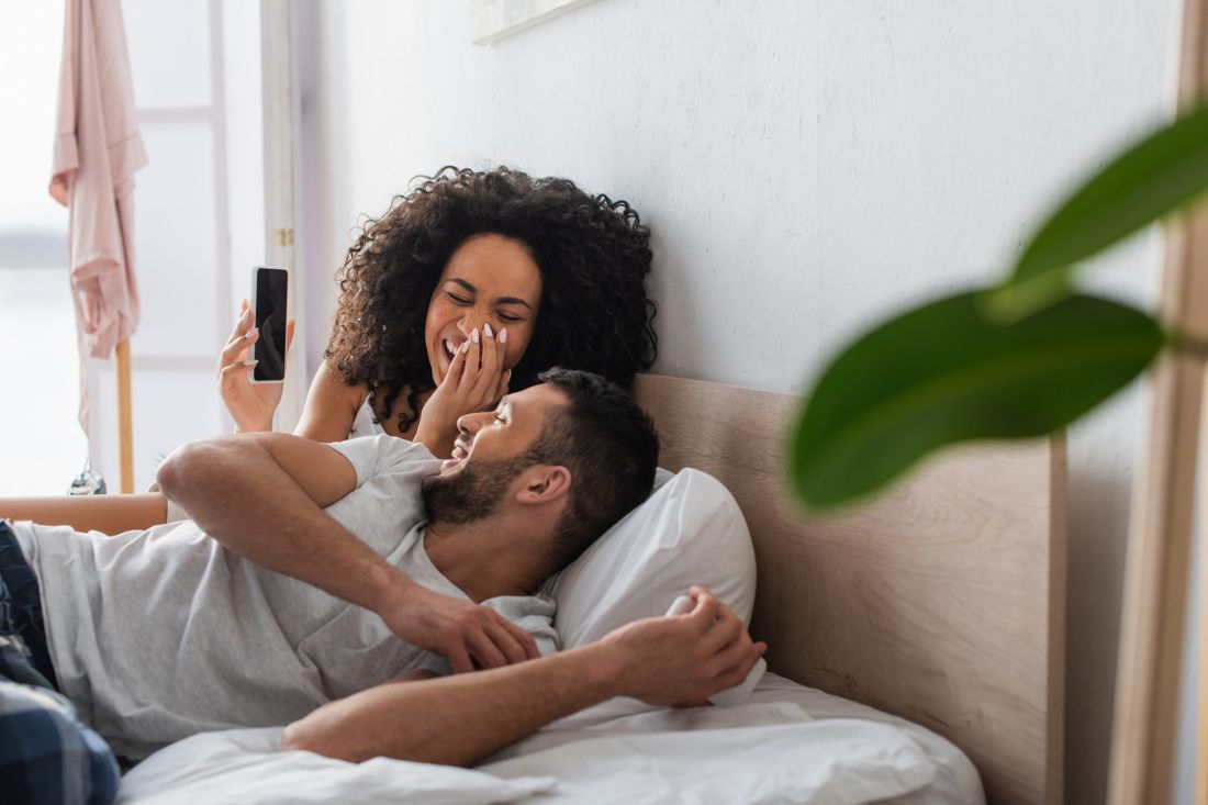 Couple laughing in bed while woman shows something through the phone.