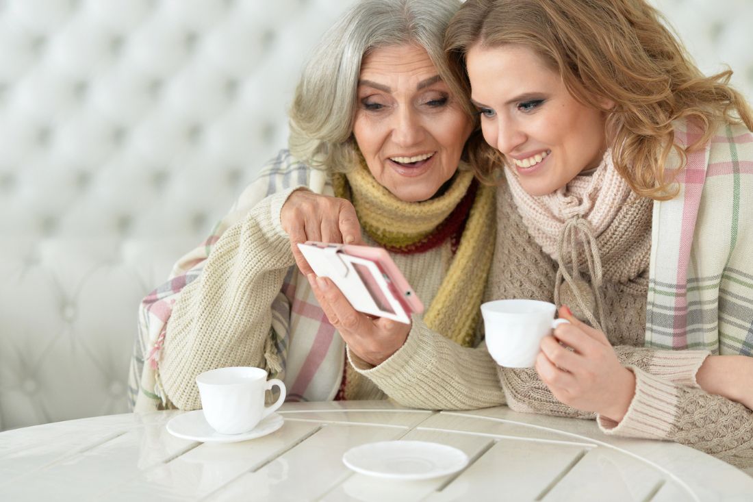 Old and young woman sitting together, having coffee and looking at a phone.