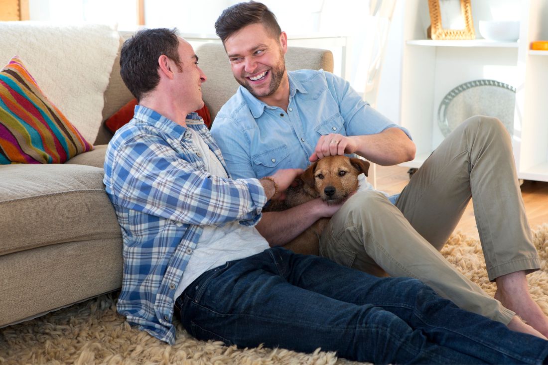 Male couple sitting and smiling with each other while holding their pet dog.