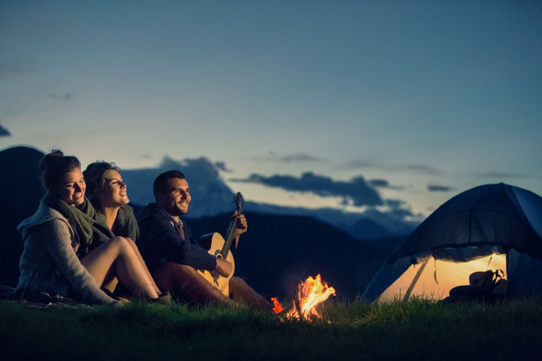 Three friends, one male and 2 females, sitting in front of a campfire on a sunset.