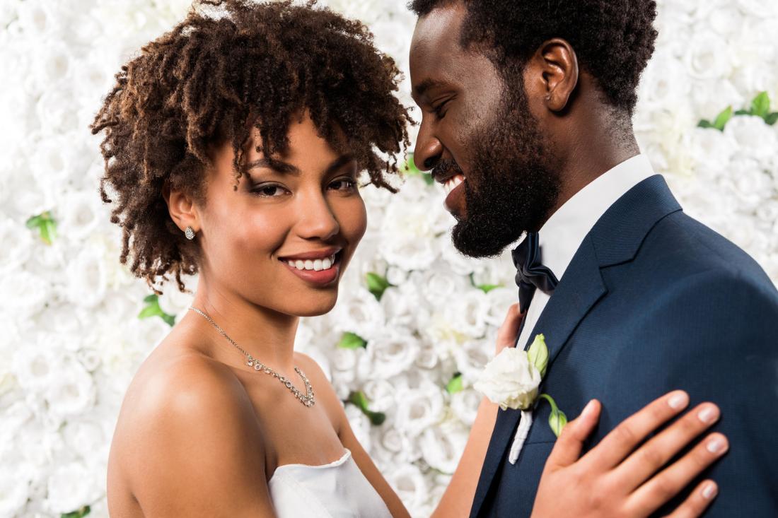 Happy African-American bride holding her smiling groom with flowers in the background.