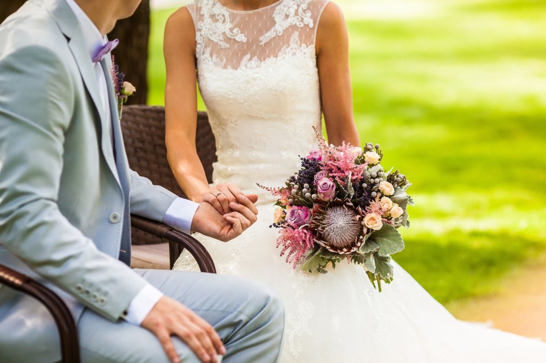Bride with bouquet and is holding the groom's hand while sitting.