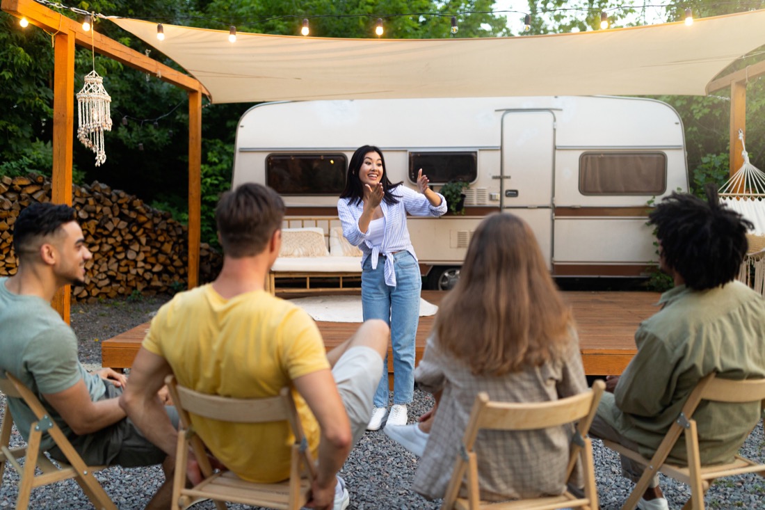 Lady playing charades to friends in front of camper trailer.