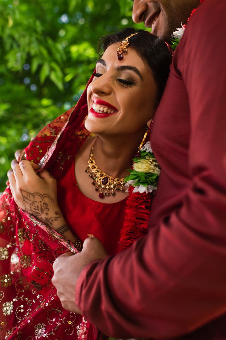 Smiling Indian bride in red dress hugging groom who is wearing a turban.