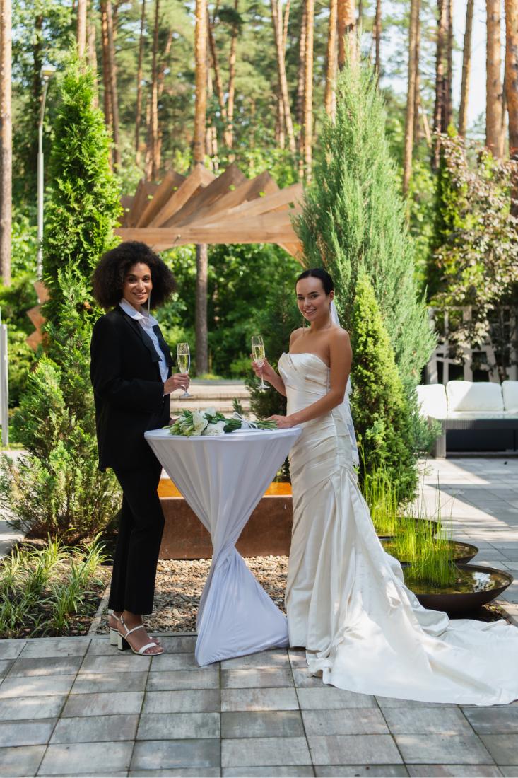 Interracial lesbian couple standing and holding wine glasses while smiling at a wedding.