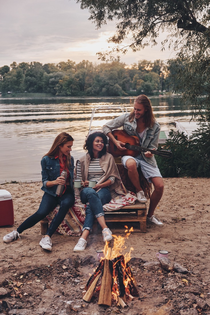 Young people in casual wear enjoying camping near lake at sunset, man playing guitar 