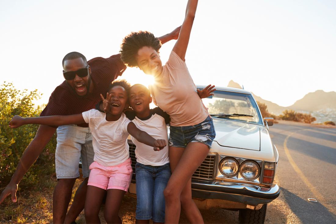 Black family of four happily posing in front of a retro car.