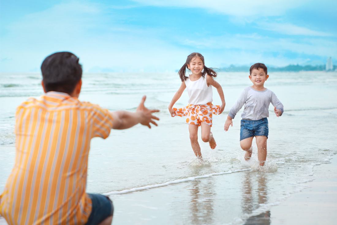 Happy young Asian boy and girl running to their dad at the beach.
