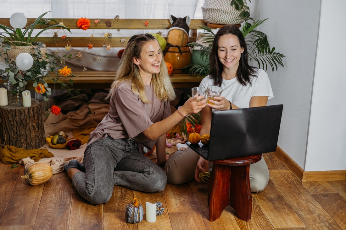 Two women cheersing drinks to laptop screen with fall decor in background.